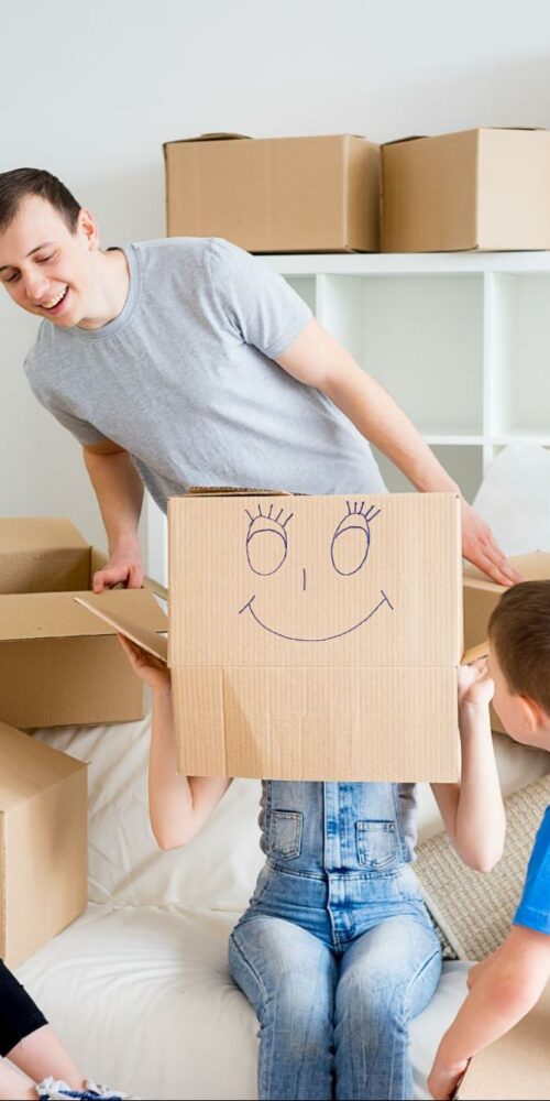 A family having fun while unpacking boxes in a bright living room, with one child holding up a box with a smiling face drawn on it.