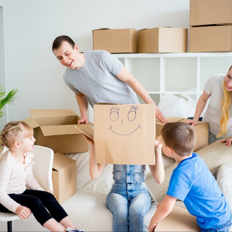 A family having fun while unpacking boxes in a bright living room, with one child holding up a box with a smiling face drawn on it.