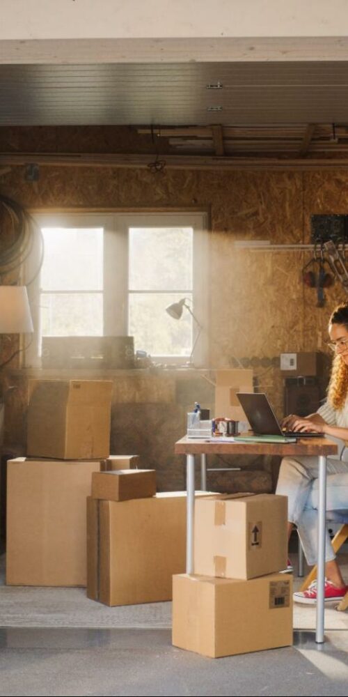 A woman sitting at a makeshift desk in a garage filled with moving boxes, working on a laptop in a sunlit space.
