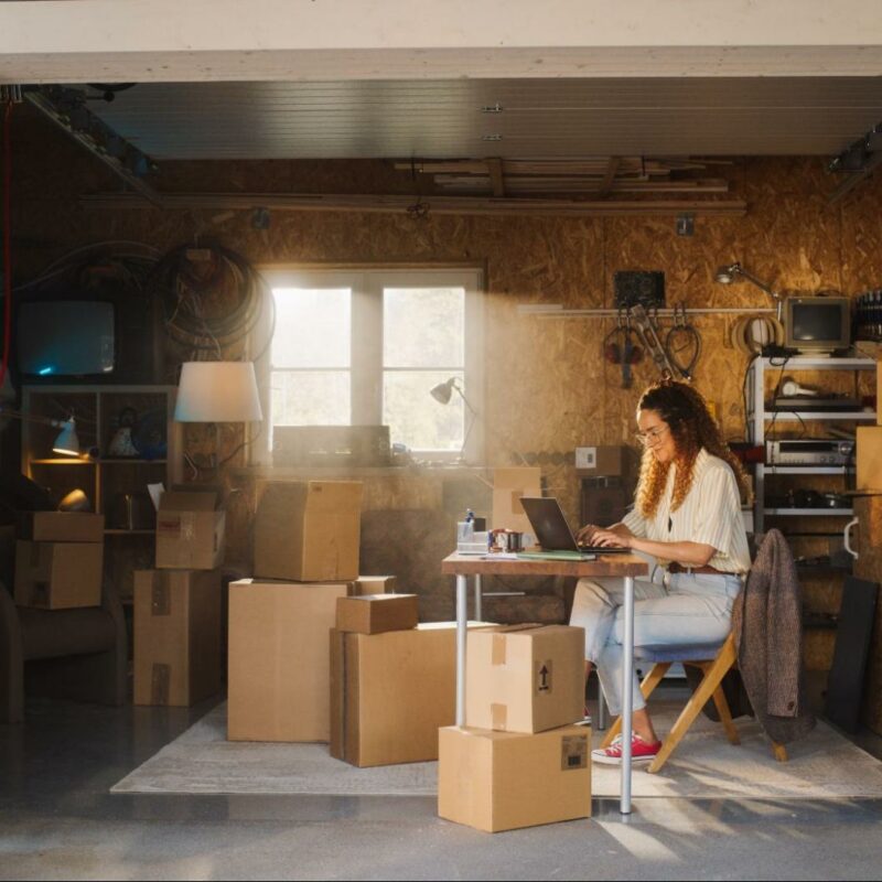 A woman sitting at a makeshift desk in a garage filled with moving boxes, working on a laptop in a sunlit space.