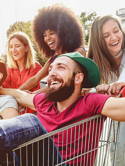 A group of young adults having fun outdoors, one of whom is sitting in a shopping cart being pushed by friends in a sunny park setting.