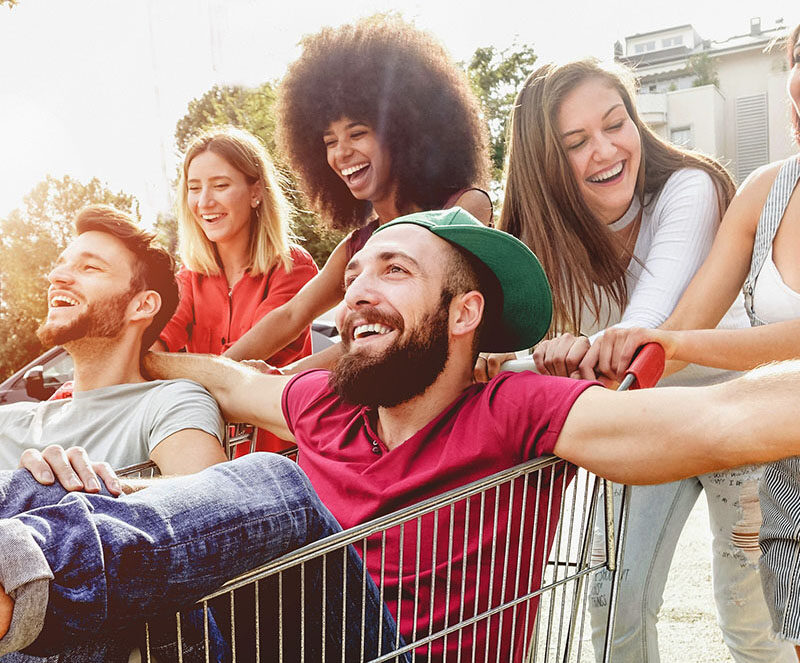 A group of young adults having fun outdoors, one of whom is sitting in a shopping cart being pushed by friends in a sunny park setting.